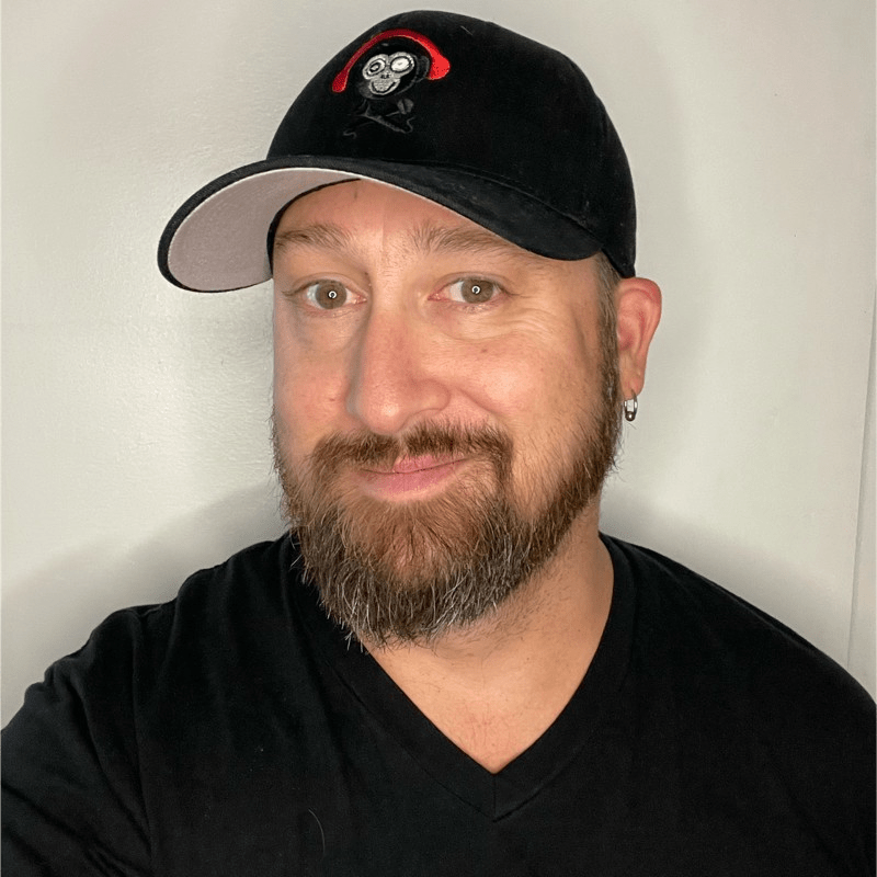 A bearded man wearing a black T-shirt and a black cap with a monkey logo, smiling slightly in front of a plain light-colored wall, appears ready to host the Try Invigor Podcast.