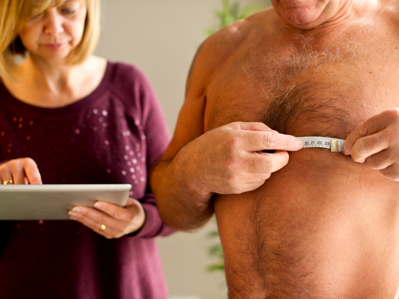 A shirtless older man measures his chest with a tape measure while a woman in a maroon sweater records information on a digital tablet nearby.