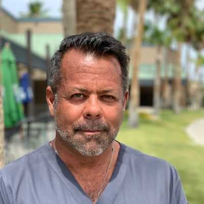 A middle-aged man with short dark hair and a beard, wearing a gray scrub top, stands outdoors with palm trees and buildings blurred in the background, representing expertise in hormones and sexual health.