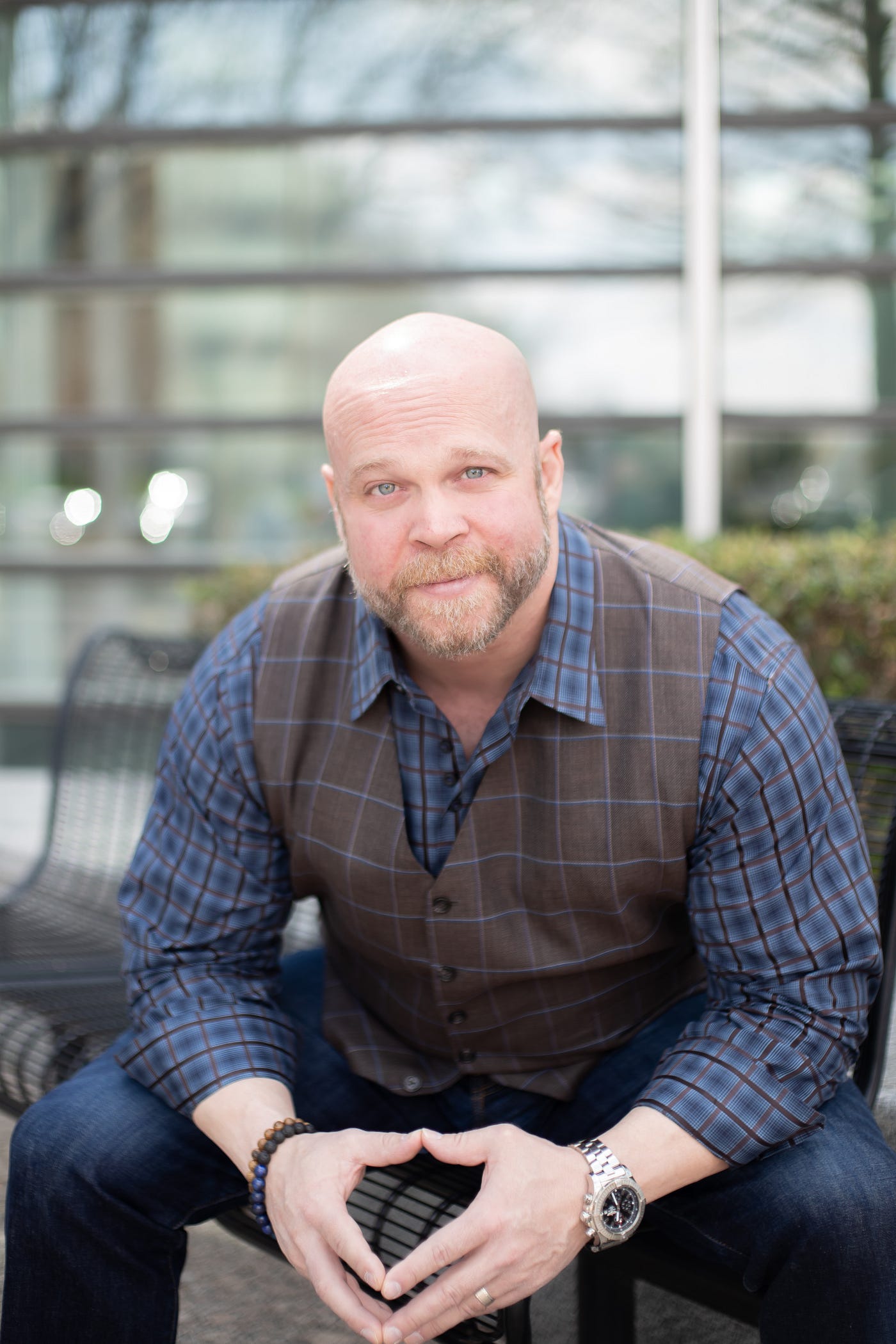 A bald, bearded man in a plaid shirt and checkered vest sits on an outdoor bench, leaning forward with hands clasped. The setting evokes a sense of holistic health, with trees and glass windows visible in the background.