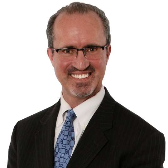 A man wearing glasses, a black pinstripe suit, white shirt, and a blue patterned tie, smiling in front of a plain white background—an advocate for gut health and healing.