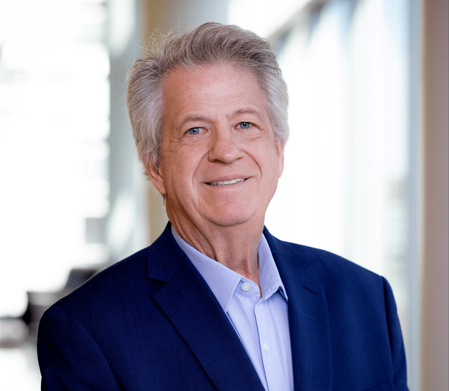 Dr. Richard Johnson, a smiling older man with gray hair, wears a dark blue suit jacket and light blue shirt, standing indoors with blurred windows and bright natural light in the background.