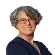 A woman with short, curly gray hair and glasses smiles confidently, embodying the spirit of Well At Work. She is wearing a dark top and standing against a plain white background.