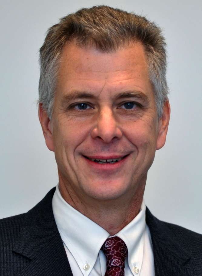 A middle-aged man with short gray hair, embodying physical intelligence, wears a dark suit jacket, white dress shirt, and patterned maroon tie, smiling confidently at the camera against a plain light background.