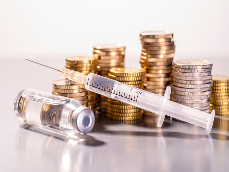 A syringe and a vial of liquid medicine are placed in front of several stacks of gold coins, symbolizing the financial aspects of healthcare or the cost of medical treatments.