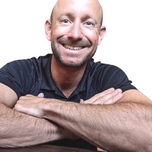 A smiling bald man with a trimmed beard, wearing a black shirt, sits at a table with his arms crossed and resting in front of him. Kyle Sela looks directly at the camera.