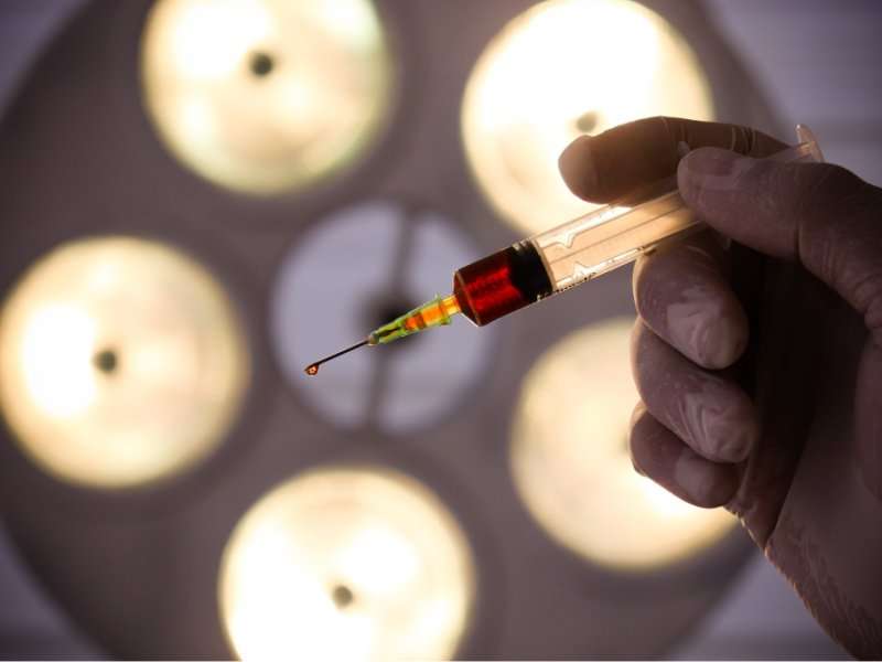 A gloved hand holds a syringe filled with liquid, with a droplet on the needle tip. Bright surgical lights shine in the blurred background, suggesting a medical or clinical setting.