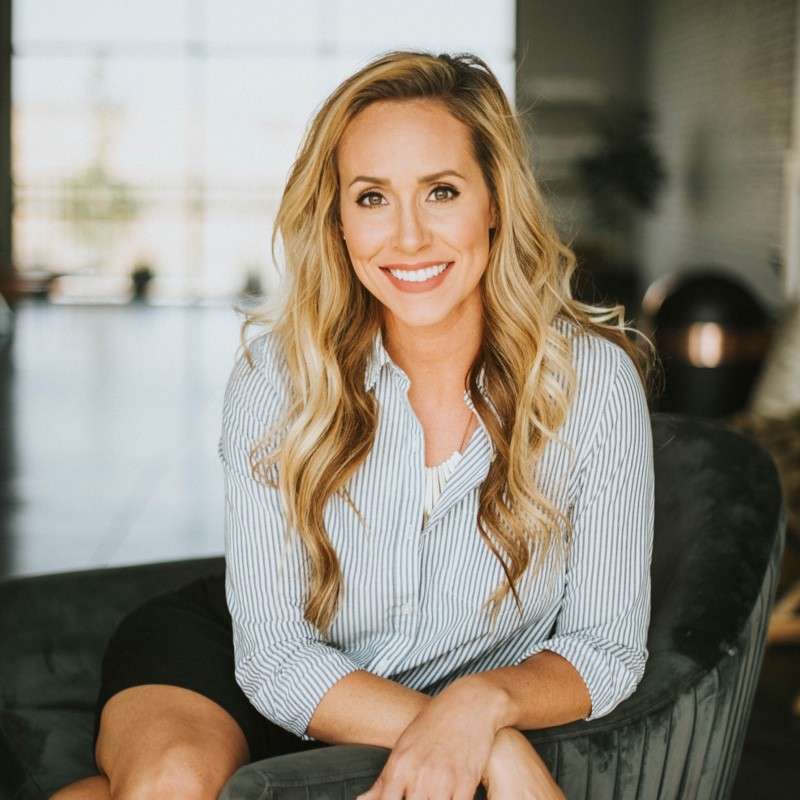 A woman with long blonde hair, wearing a light blue striped shirt, sits on a dark cushioned chair and smiles at the camera in a bright, modern room—radiating well-being inspired by Yoga for Longevity.