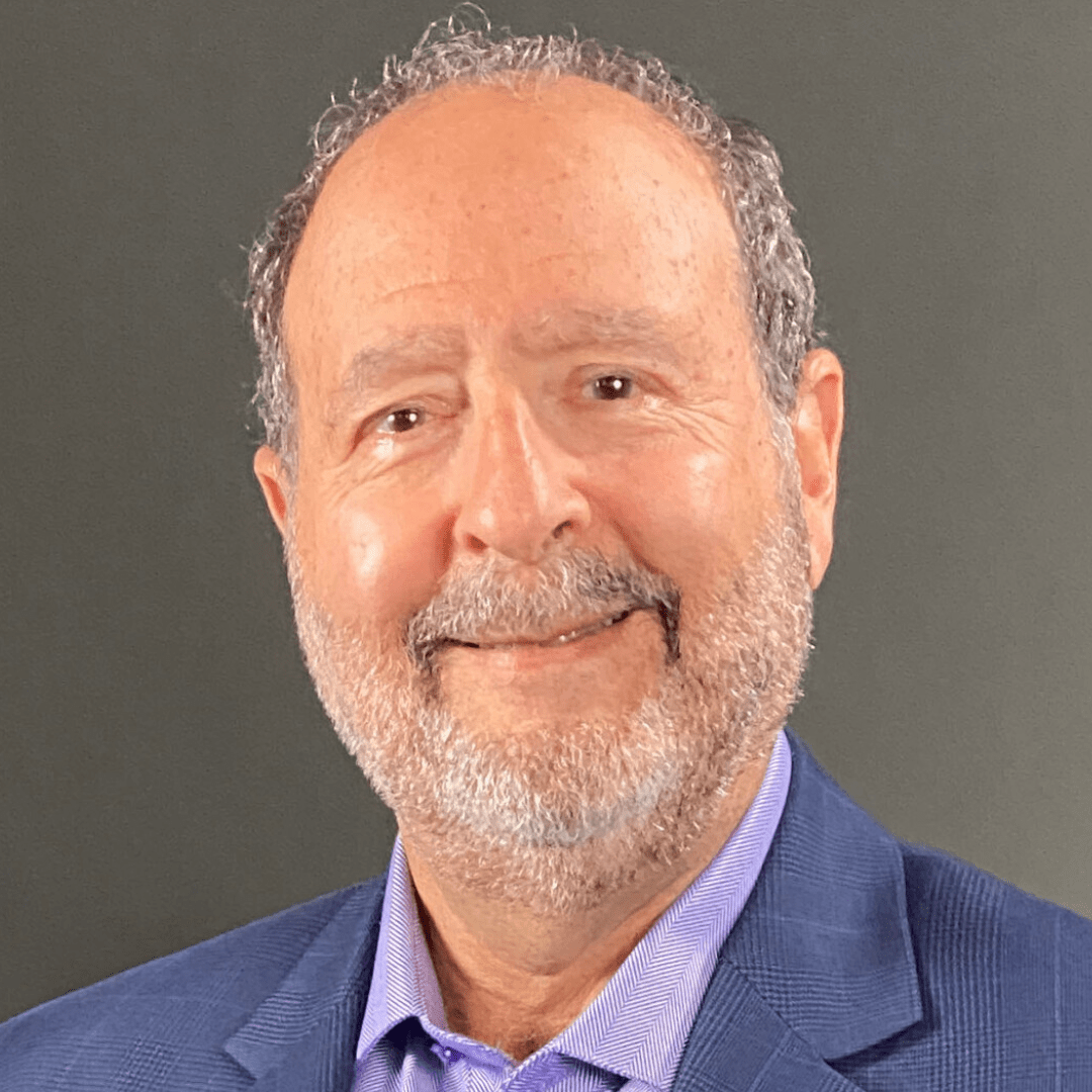 A smiling older man with short curly gray hair, a full beard, and mustache, wears a blue jacket over a light purple collared shirt while posing in front of a plain gray background, embodying the vitality found in The Power of 5.
