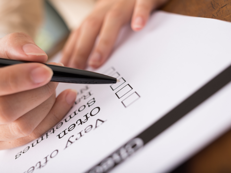 A hand holding a pen is about to check a box on a paper survey or questionnaire. The text on the form includes response options like Very Often and Sometimes.