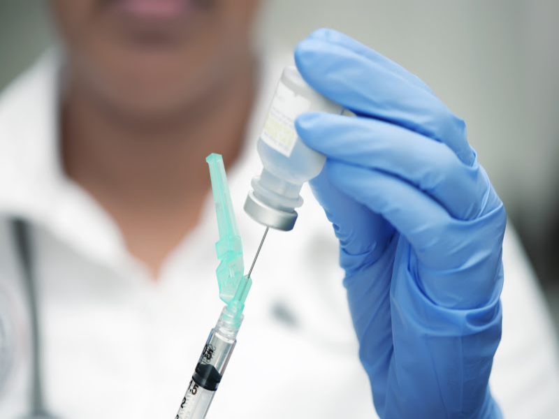 A person wearing blue gloves fills a syringe with liquid from a vial, preparing for an injection. The background is blurred, focusing attention on the syringe and vial.