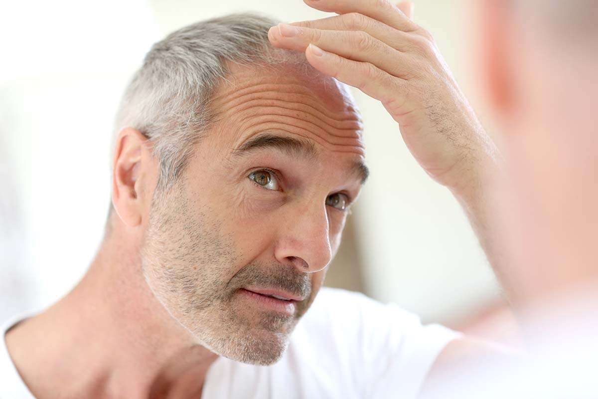 A middle-aged man with gray hair and a beard looks at his reflection in the mirror, lifting his hair and examining his hairline with a concerned expression, perhaps wondering how long it takes for finasteride to work.