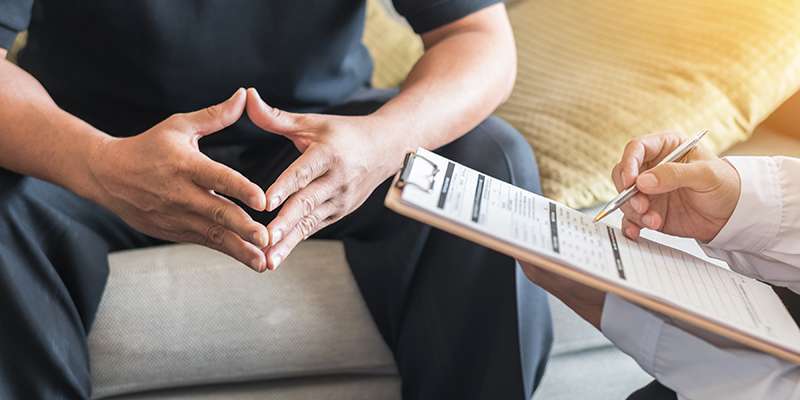 A person sits on a couch with hands clasped, while another person holding a clipboard and pen takes notes, suggesting a counseling or therapy session.