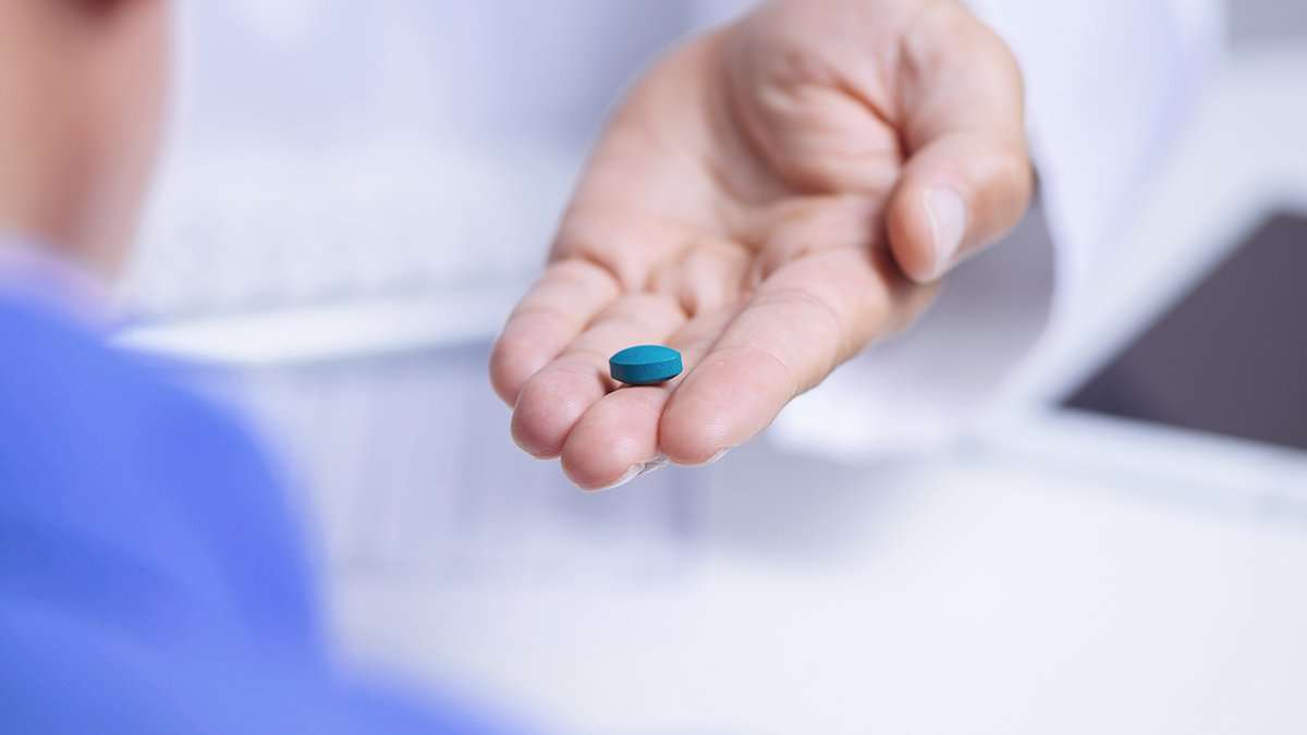A person’s hand holding out a single teal-colored pill toward another person in a blue shirt, suggesting the offering or administration of medication in a clinical setting.