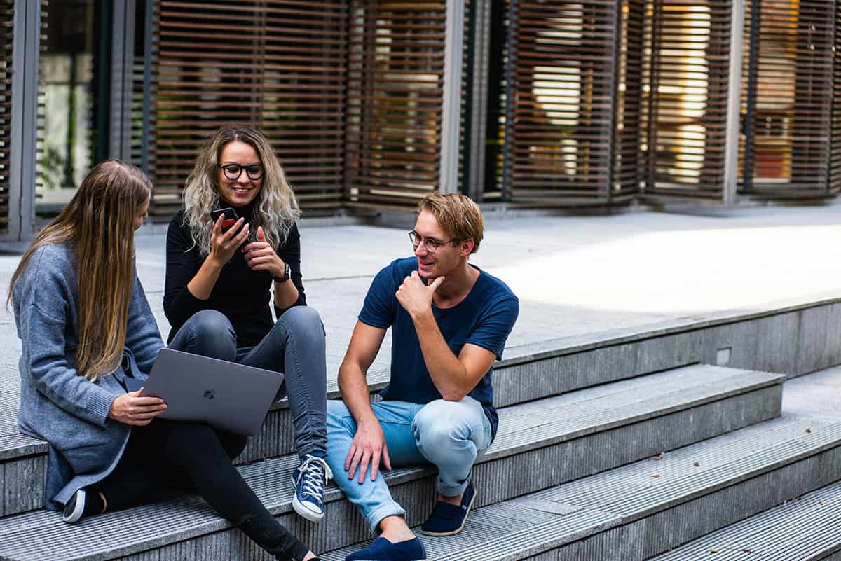 Two women and a man sit at the steps of a building, laughing while holding cellphones and a laptop.